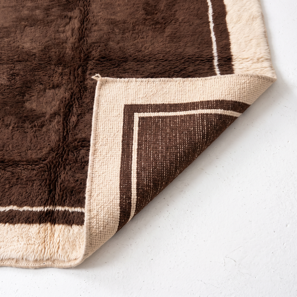 Corner of a chocolate brown Moroccan rug folded back to show the flat hand-knotted back, which mirrors the cream border, beside the shaggy wool pile
