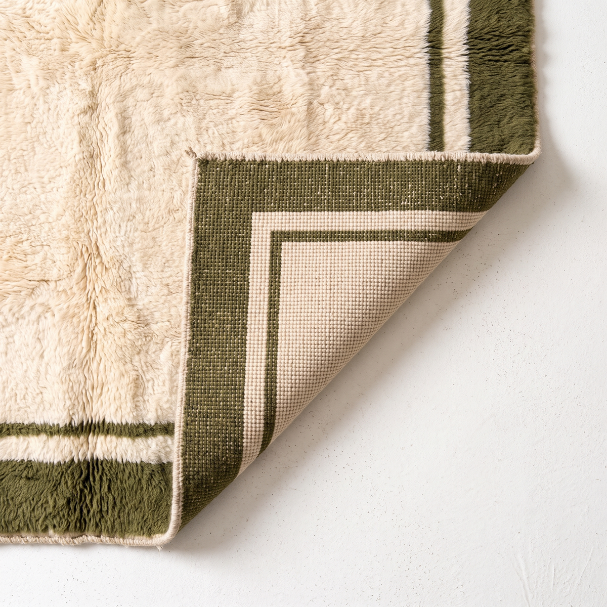 Corner of a cream Moroccan rug folded back to show the flat hand-knotted back, which mirrors the olive green border, beside the shaggy wool pile